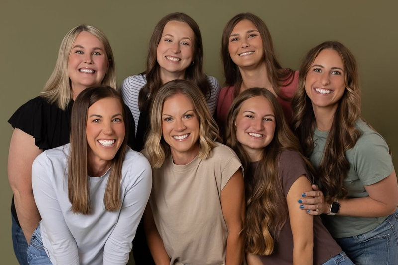 Female dental hygienists and administrative team members smiling together in front of an olive green studio backdrop