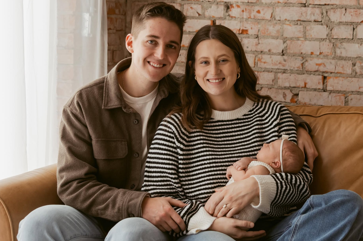 Young couple sitting on a leather couch against an exposed brick wall, holding their newborn baby.